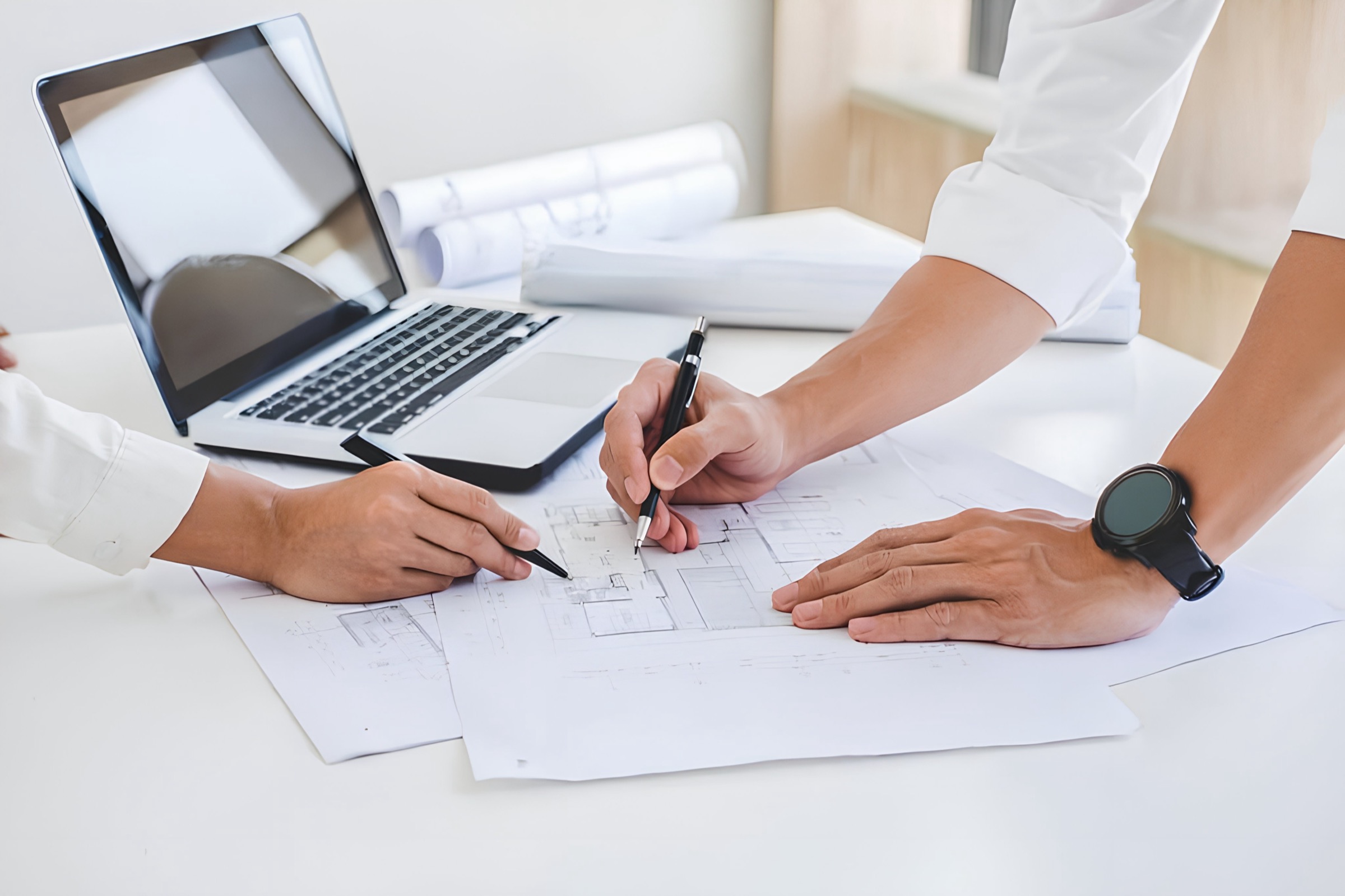 Two people reviewing construction blueprints on a bright desk with a laptop